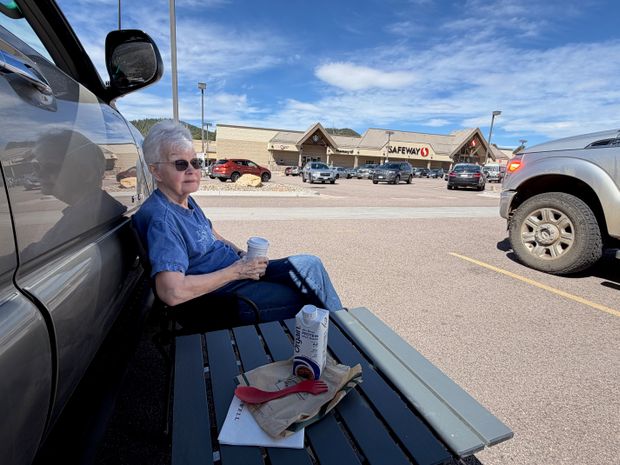 A woman resting with coffee beside a vehicle in a Woodland Park parking lot