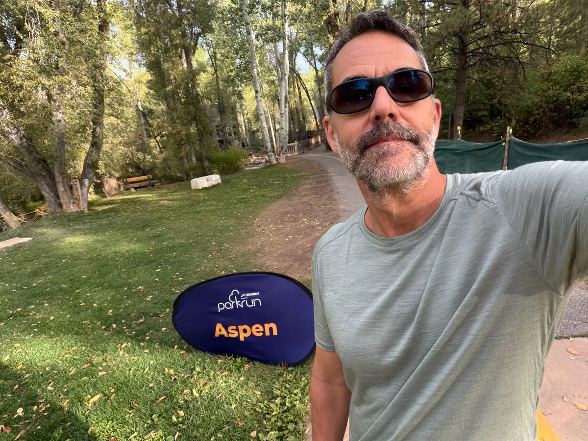 A runner taking a selfie beside the Aspen parkrun sign on a wooded course